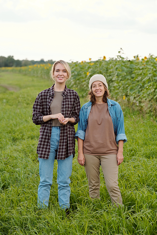 Two cheerful female farmers in workwear looking at you with smiles while standing in front of camera against green field and country road