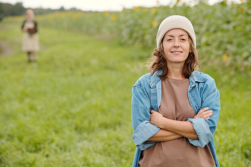 Mature cheerful female farmer in beanie and workwear looking at you with smile while standing in front of camera against green field and sky