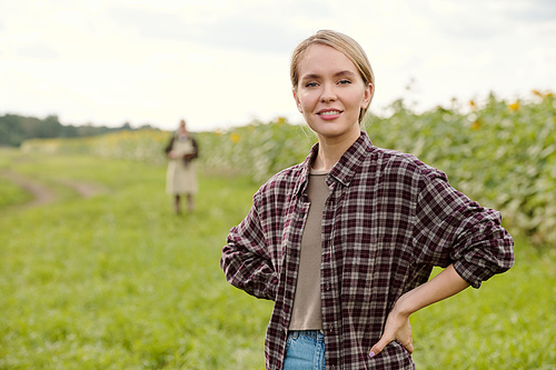 Young blond smiling female farmer in workwear looking at you while standing in front of camera and using digital tablet against green field