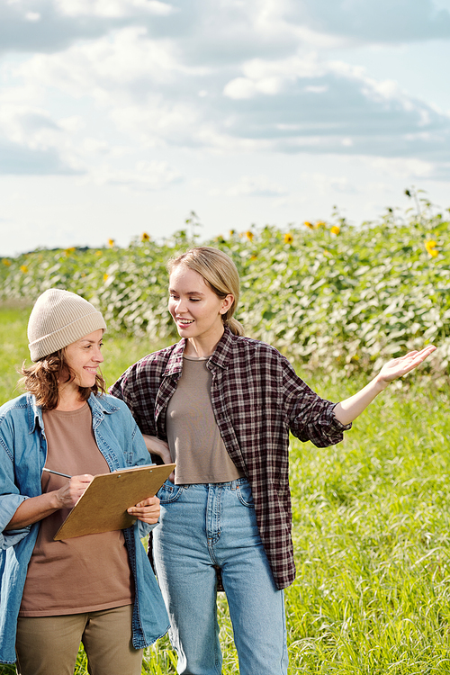 Two cheerful female farmers in workwear looking at sunflower field in front of camera while one of them making notes during discussion