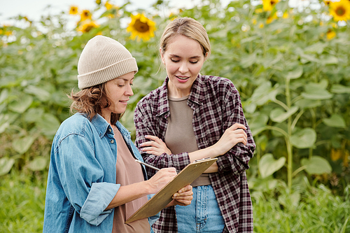 Two cheerful female farmers in workwear looking at sunflower field in front of camera while one of them making notes during discussion