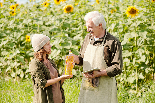 Happy senior male farmer in workwear passing bottle of sunflower oil to his mature female colleague against green field on sunny summer day