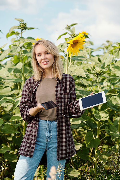 Young blond smiling female farmer in workwear looking at you while standing in front of camera and using digital tablet against green field