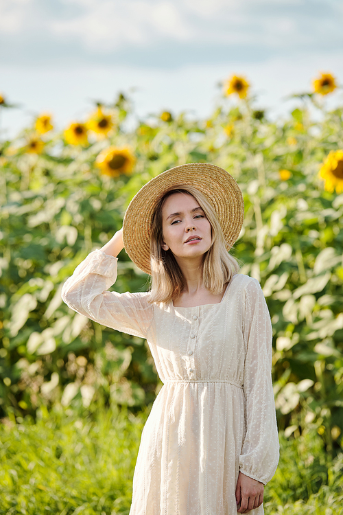 Happy young blond woman in hat and white romantic dress standing on green grass in front of camera against sunflower field on sunny day