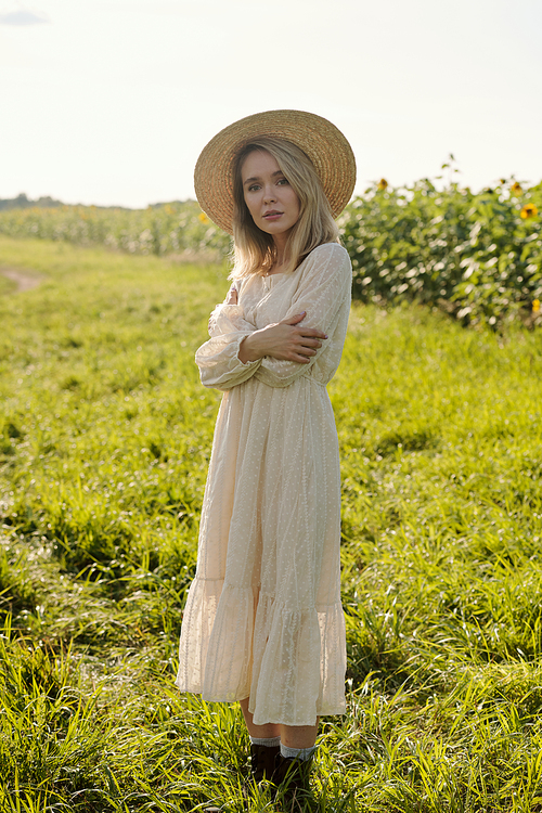 Gorgeous young blond woman in straw hat and white romantic dress standing in front of camera against sunflower field on sunny day