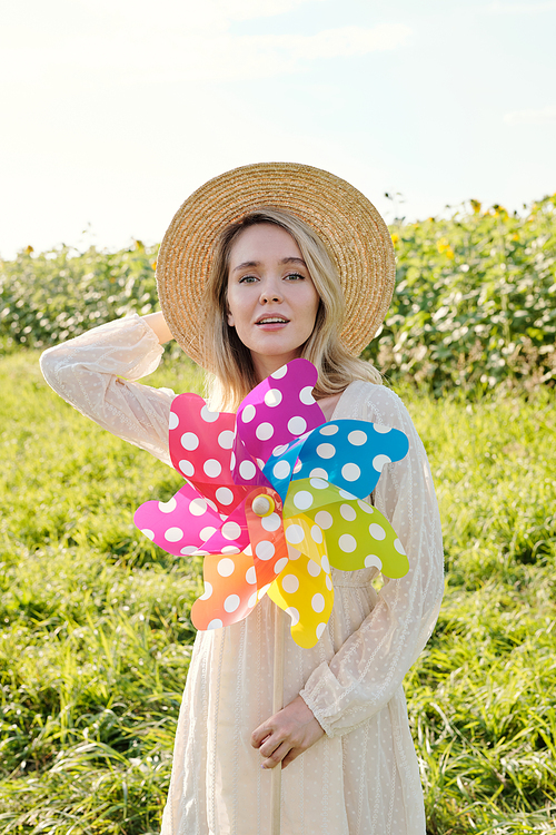 Gorgeous young blond woman in hat and country style dress holding large polkadot whirligig while standing against green grass and sunflower field