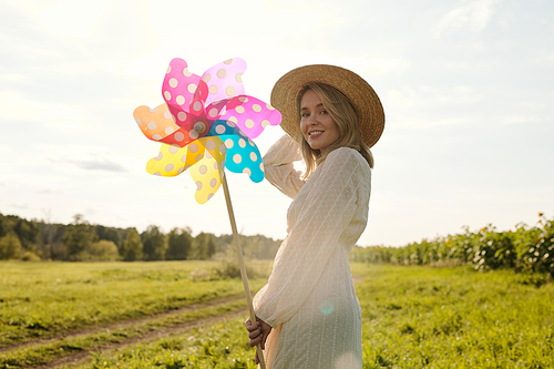 Back view of young elegant woman in hat and country style dress holding large polkadot whirligig while standing in front of green grass and road