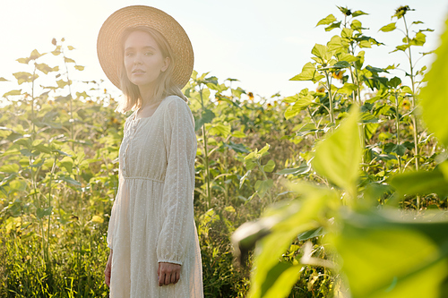 Beautiful young woman in straw hat and white country style dress looking at you while standing in front of camera against sunflowers at sunset