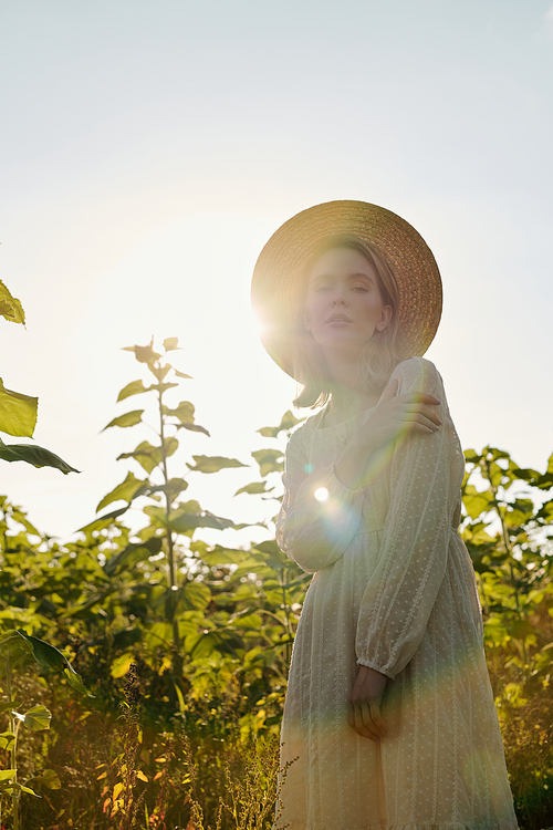 Beautiful young blond woman in straw hat and white country style dress standing in front of camera against sunflower field over cloudy sky