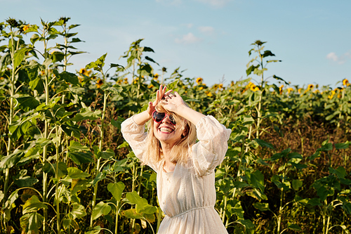 Blond young woman in sunglasses and white country style dress standing in front of camera against sunflower field and blue sky in summer