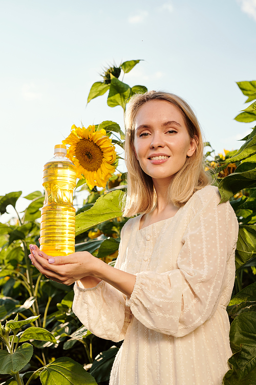 Gorgeous blond young woman holding bottle of sunflower oil while standing against large flowers in front of camera in the field