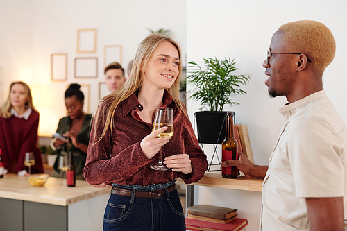 Happy young brunette woman in pink pullover clinking flute of champagne with that of her boyfriend while enjoying home party with friends