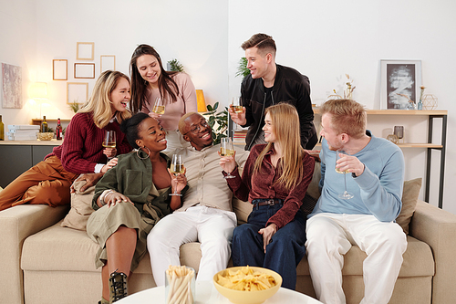 Group of young cheerful intercultural friends in smart casualwear sitting on large couch in living-room in front of camera and toasting