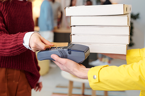 Young smiling blond female in smart casualwear holding credit card over screen of payment machine in hand of male courier in yellow uniform