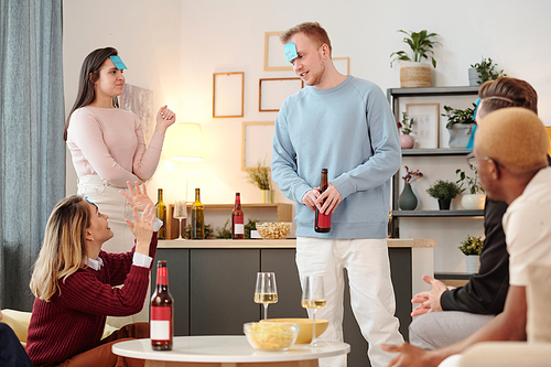 Group of young cheerful intercultural friends in smart casualwear sitting by small white round table in living-room, having pizza and drinks