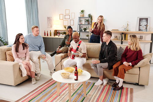 Group of young intercultural friends sitting on couches around small table with snacks and drinks and listening to singing of African guy at party