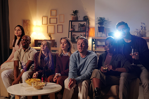 Group of young intercultural friends sitting on long couch by small table with snacks and watching movie together in living-room at party