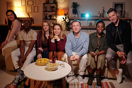 Group of young intercultural friends relaxing on couch in living-room and watching interesting movie together while enjoying home party