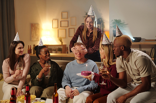 Cheerful young intercultural friends congratulating excited guy on his birthday looking at burning candle on glazed delicious cake