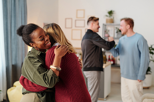 Happy young African woman in smart casualwear giving hug to guy in blue pullover in front of camera against their friends embracing and talking