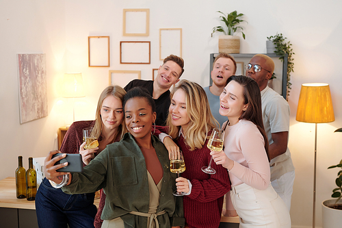 Large group of happy young intercultural friends in smart casualwear making selfie while cheering up with flutes of champagne at party