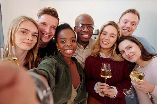 Large group of happy young intercultural friends in smart casualwear making selfie while cheering up with flutes of champagne at party