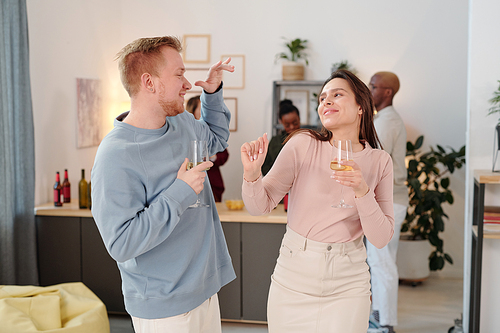 Happy young intercultural couple with bottles of beer looking at you while enjoying home party against their friends chatting and having drinks