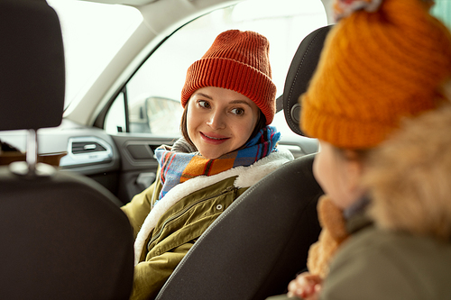Happy young mother in warm winterwear looking at her little daughter sitting on backseat of car while travelling to the country in winter
