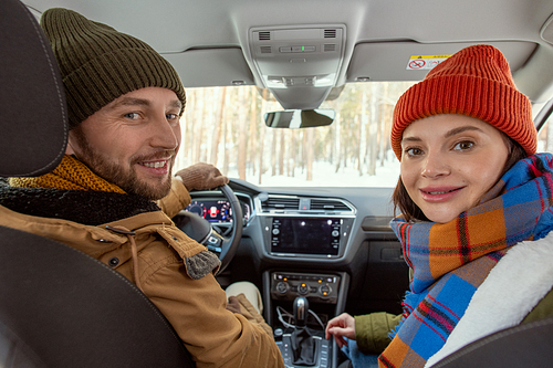 Young cheerful couple in warm winterwear looking at you with smiles while sitting in car and traveling to their countryhouse, man holding by steer