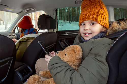 Cute cheerful little girl in warm winterwear looking at you with toothy smile while sitting in the backseat of car and traveling with her parents