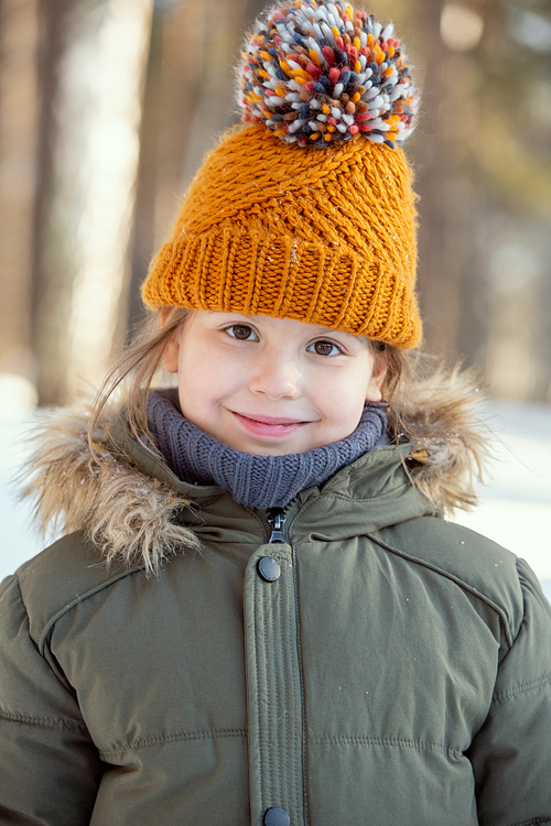 Adorable child in warm jacket and knitted brown beanie looking at you with smile while standing in front of camera in natural environment