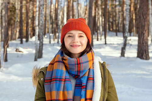 Young cheerful woman in warm jacket, checkered scarf and beanie looking at you with smile while standing in front of camera in natural environment