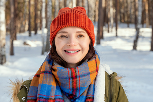 Young female in warm jacket, checkered scarf and beanie looking at you with toothy smile while standing in front of camera in natural environment