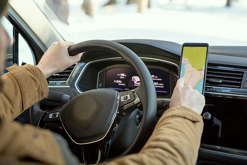 Hands of young man in warm winterwear holding by steer while sitting in car and pointing at map on screen of smartphone during travel