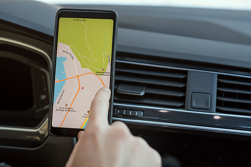 Hand of young man sitting in car and pointing at map on screen of smartphone while choosing place where he going to travel