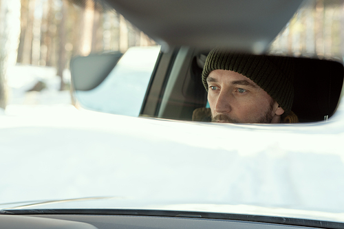 Reflection of face of young active man in winterwear looking through front window of car while driving to country house or some other place