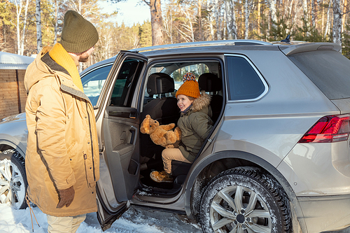 Young father opening door of car for his adorable little daughter in winterwear sitting on backseat while traveling to the country