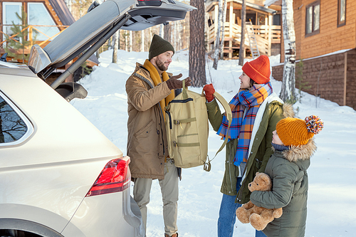 Young family of three in warm winterwear taking their buggage out of trunk while standing by back of the car during winter travel