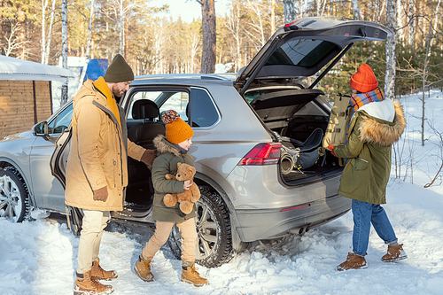 Happy cute little girl with teddybear coming up to mother taking rucksack with baggage out of car trunk on arrival in their country house