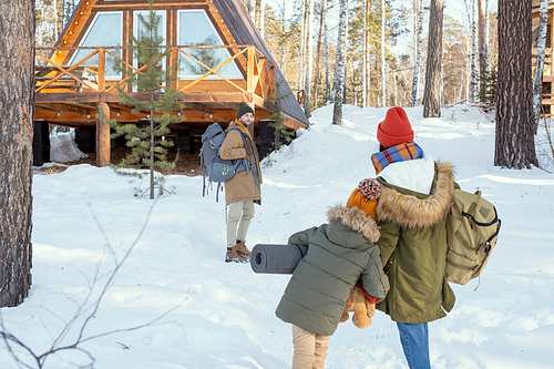 Young man with big rucksack standing in the forest against country house and looking at his wife and daughter moving towards him