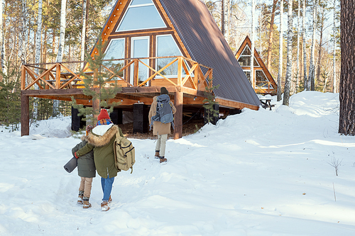 Young man with big rucksack moving towards country house while his wife and their little daughter in warm winterwear following him