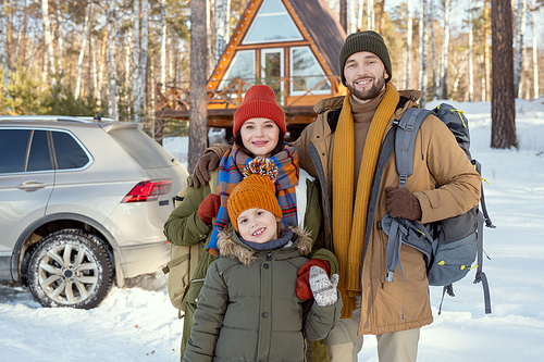 Happy young family of three in warm winterwear standing in front of camera against their country house and looking at you outside