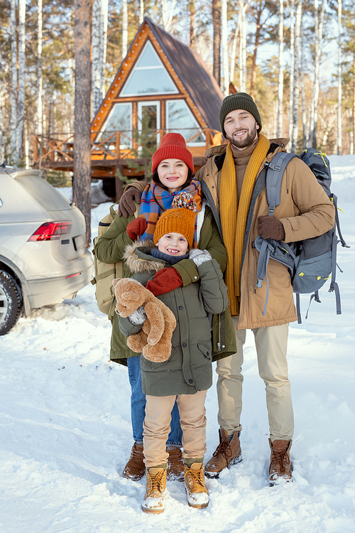 Young affectionate family of three in warm winterwear standing in front of camera against their country house and looking at you outside