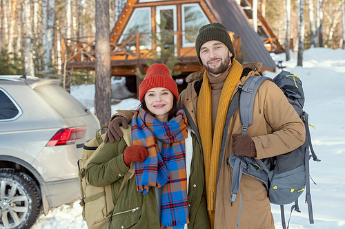 Young affectionate couple in warm winterwear standing in front of camera against their car and country house and looking at you outside