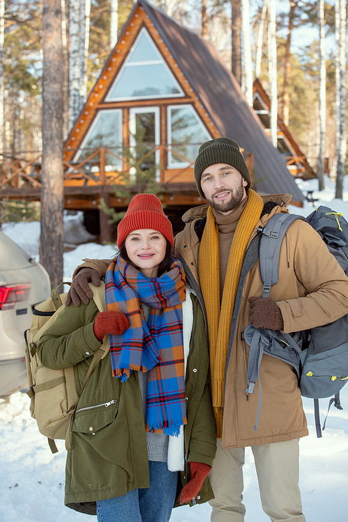 Young affectionate couple with big backpacks standing in front of camera against their car and country house and looking at you outside