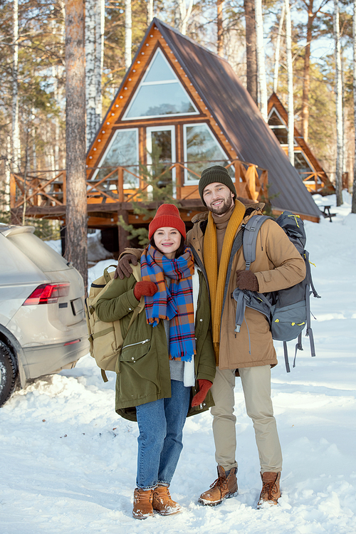 Young cheerful couple with backpacks standing in front of camera against their car and country house and looking at you on winter weekend