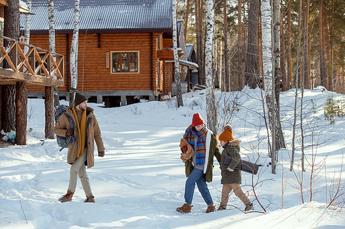 Young man with rucksack moving towards country house in the forest while his wife and their little daughter in warm winterwear following him