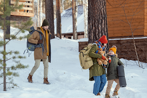 Young man with rucksack following his wife and their little daughter while moving towards country house on arrival in modern winter resort