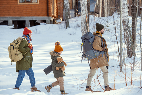 Young man with rucksack, his wife and their cute little daughter with teddybear moving towards country house in the forest on winter day