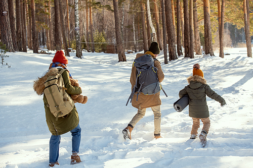 Father and little daughter in warm winterwear having fun in snowdrift while moving towards country house, mother with rucksack following them